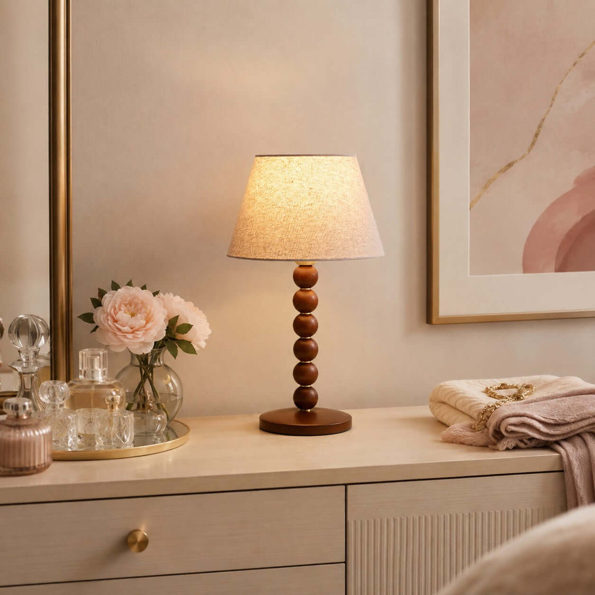 Lit Clérachic bead-column lamp on a cream dresser with pink roses, perfume bottles and a folded blanket beside it.