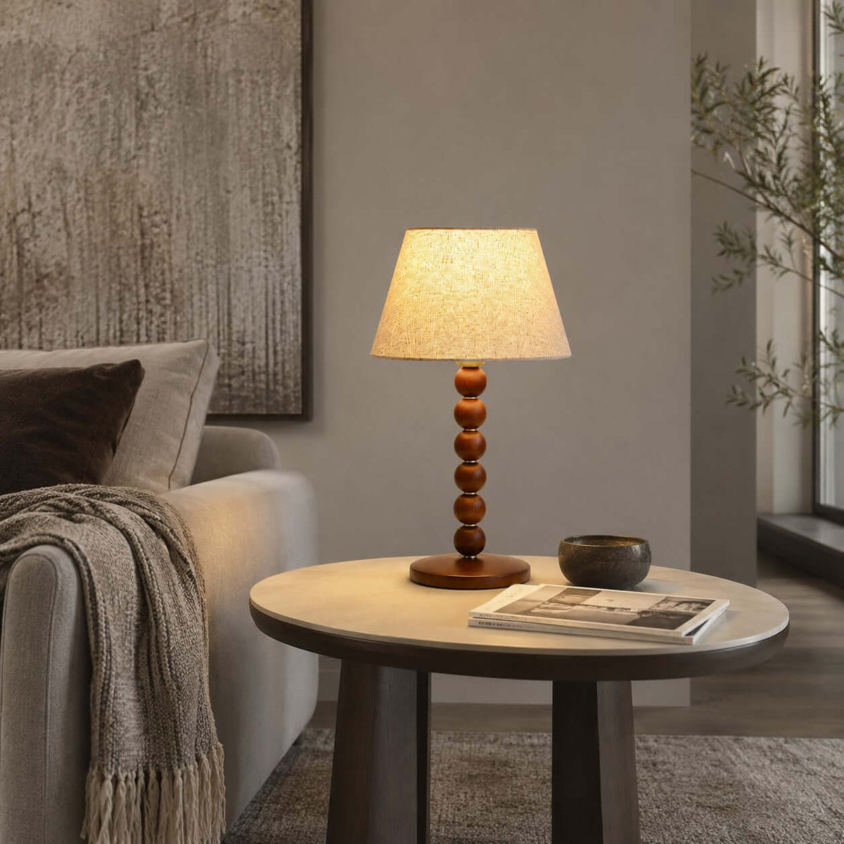 Beside a grey sofa, a Clérachic bead-base lamp stands on a round table with a magazine and a small dark bowl.