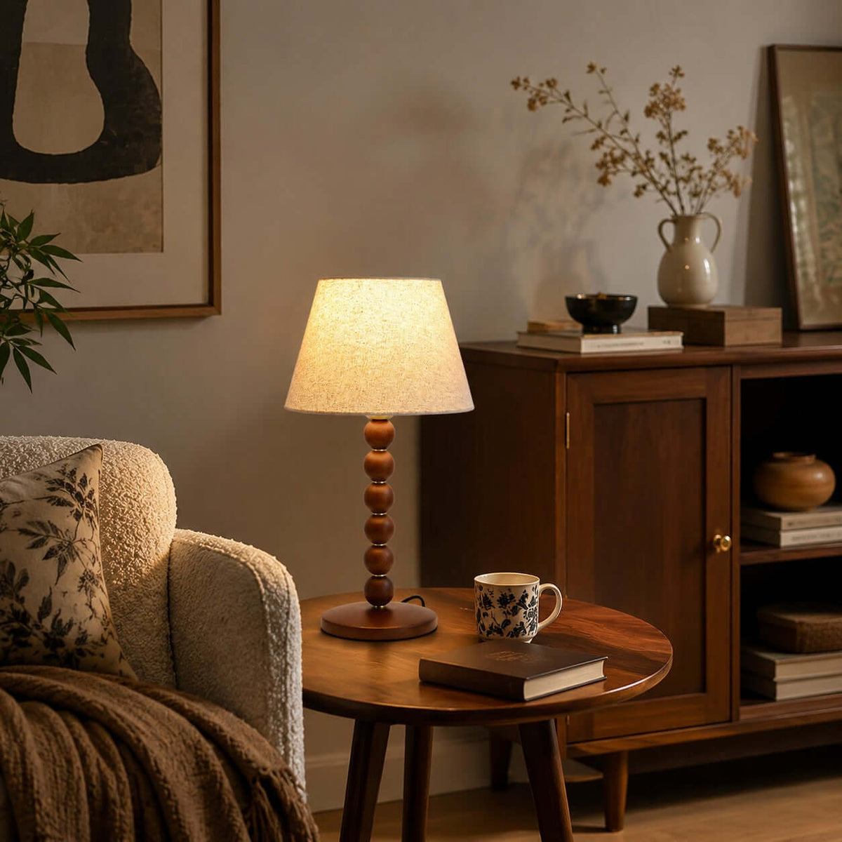 A Clérachic table lamp on a small round wooden table, next to a boucle armchair, a mug of tea and a closed book.