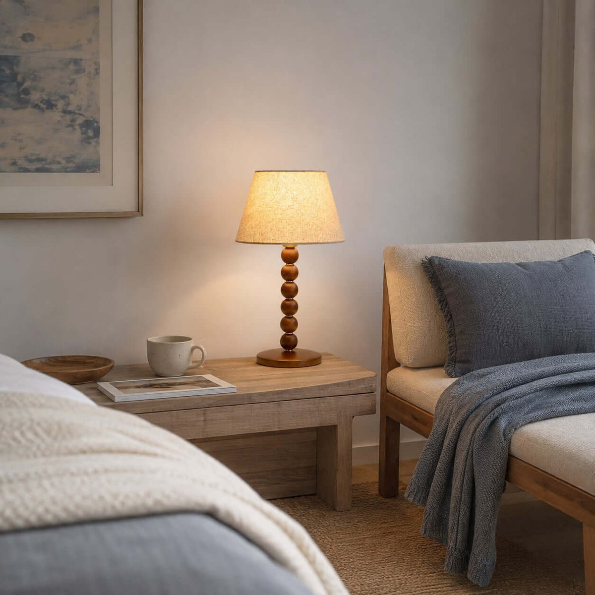 Clérachic lamp glowing on a pale wooden bedside table with a mug and a magazine, beside a daybed with a blue throw.