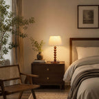 A Clérachic table lamp on a two-drawer walnut nightstand next to a bed, with a rattan chair and a small potted plant nearby.