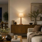 Evening view of this Clérachic lamp on a low wooden sideboard in a living room, with books, a vase of branches and a cream sofa.