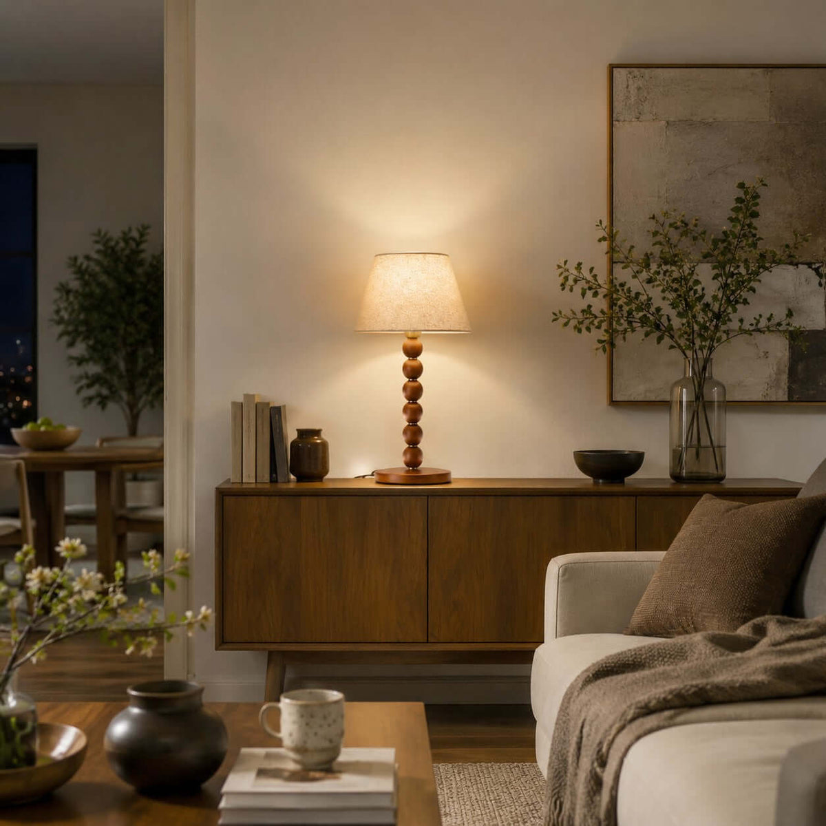 Evening view of this Clérachic lamp on a low wooden sideboard in a living room, with books, a vase of branches and a cream sofa.