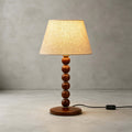 The walnut-finish beaded stem, round wooden base and beige fabric shade of this Clérachic lamp, on a plain concrete-grey floor.