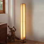 Lit Clérachic floor lamp with a beige pleated shade in a room corner, beside a chest of drawers and a driftwood piece.