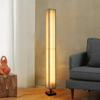 Beige pleated Clérachic floor lamp glowing warm beside a grey armchair, with a wooden block and a sculpture nearby.