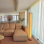 Tall Clérachic floor lamp glowing at the end of a sofa, next to a wall of sheer curtains in a large living room.
