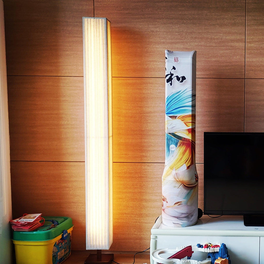 Lit Clérachic floor lamp against a warm wood-panelled wall beside a television stand, with toys in a box below.