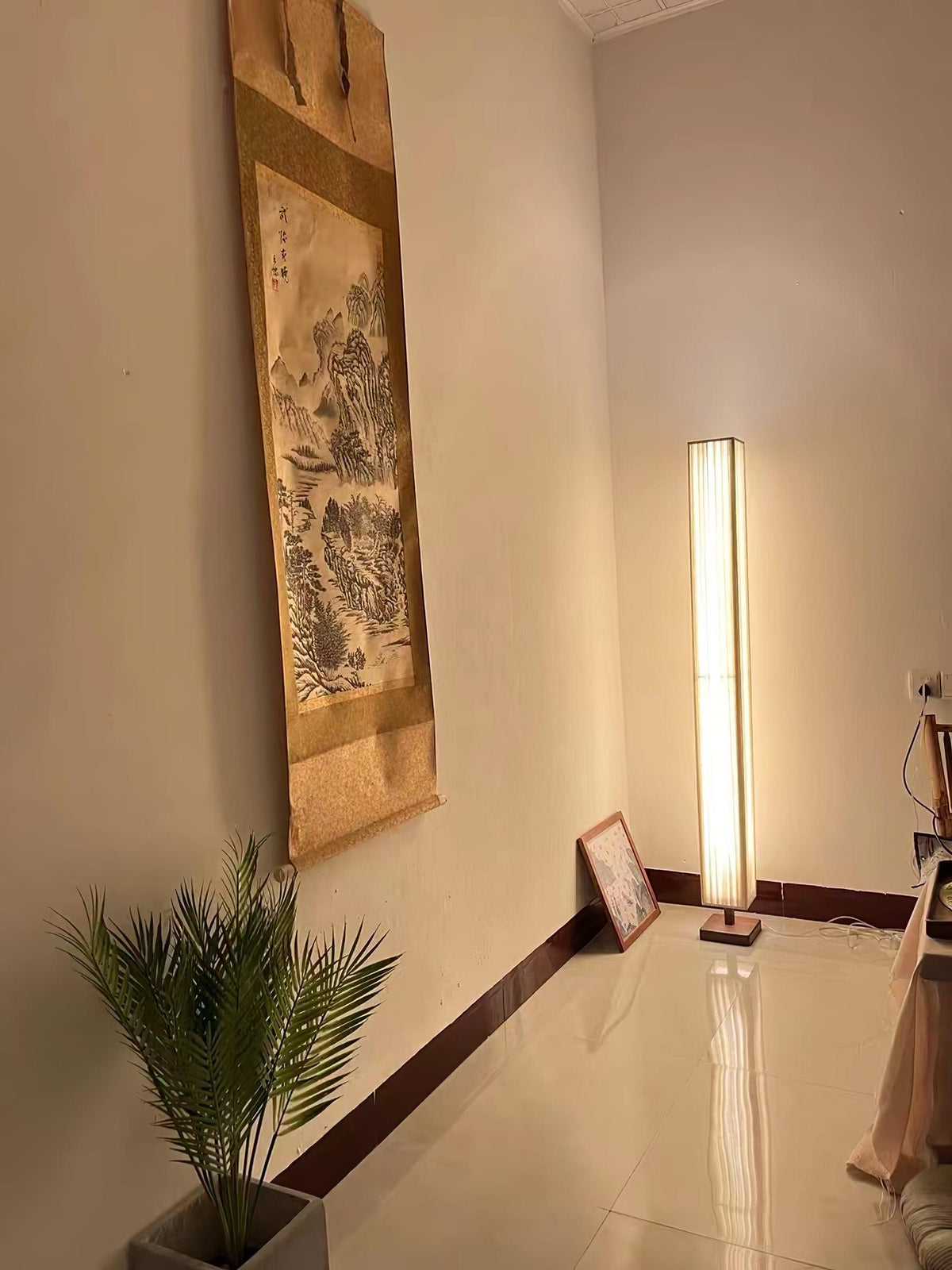 A Clérachic floor lamp glowing in a corner of a polished-floor room, with a hanging scroll and a potted palm.