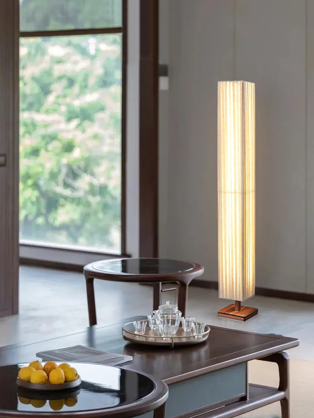 Lit Clérachic floor lamp by a large window, behind a dark coffee table with a tea set and a bowl of lemons.