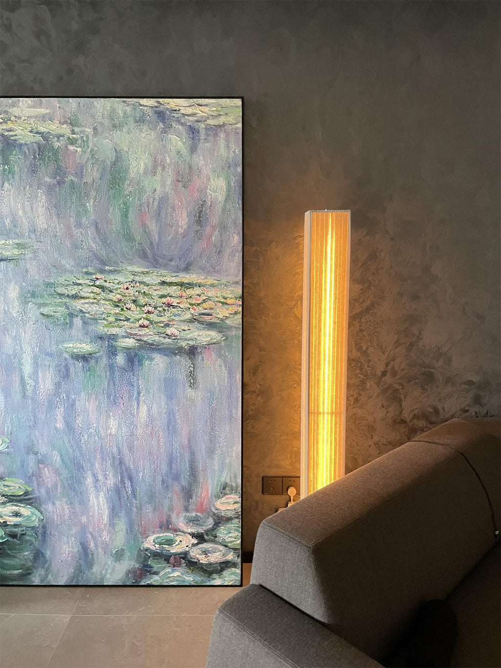 Warm light from a Clérachic floor lamp beside a large water-lily painting, with the arm of a grey sofa in front.