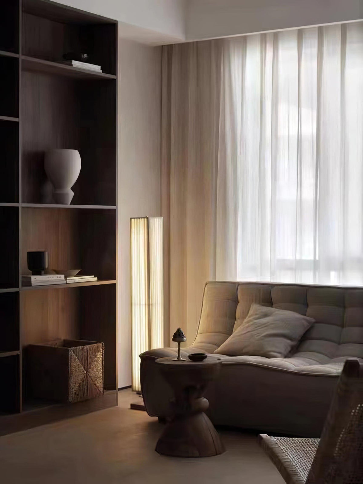 Clérachic floor lamp lit in the corner of a living room, beside a dark bookcase, a wide sofa and a sheer curtain.