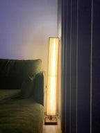Pleated Clérachic floor lamp glowing in a dim room, between a dark green sofa and navy curtains.