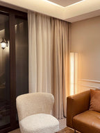 A Clérachic floor lamp lit softly behind a cream armchair and a tan leather sofa, with long curtains at the window.