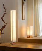Clérachic floor lamp beside a driftwood branch, with a smaller lit table lamp on a raised wooden platform behind.