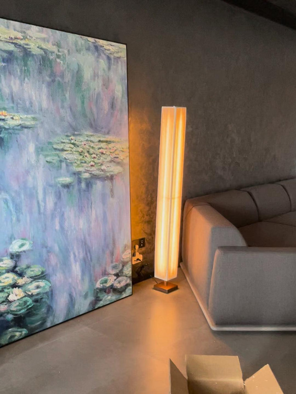 Clérachic floor lamp glowing warm in front of a large water-lily painting, with a grey sofa and a cardboard box nearby.