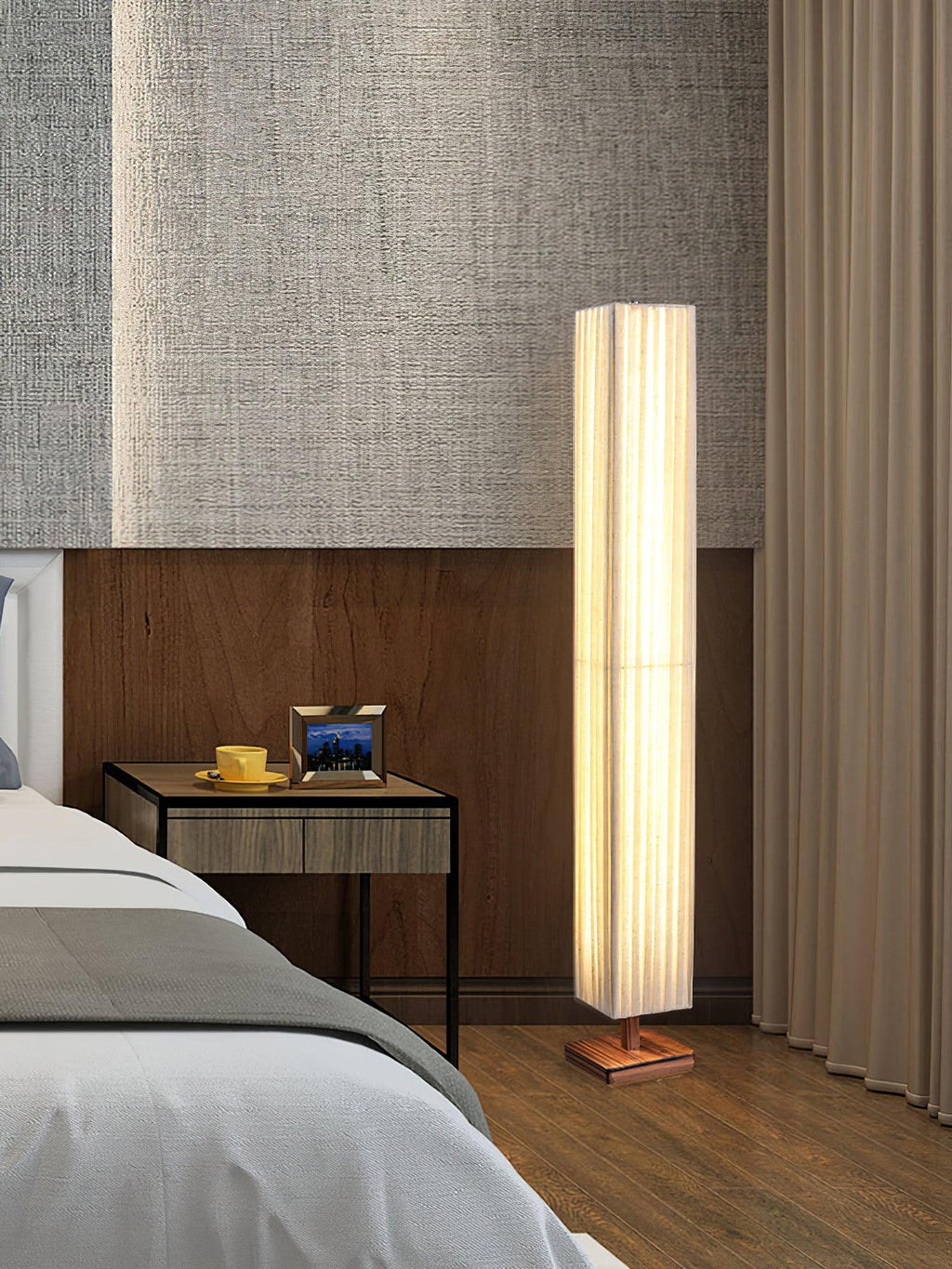 Lit Clérachic floor lamp at the foot of a bed, beside a bedside table with a cup and a framed photo, and long curtains.
