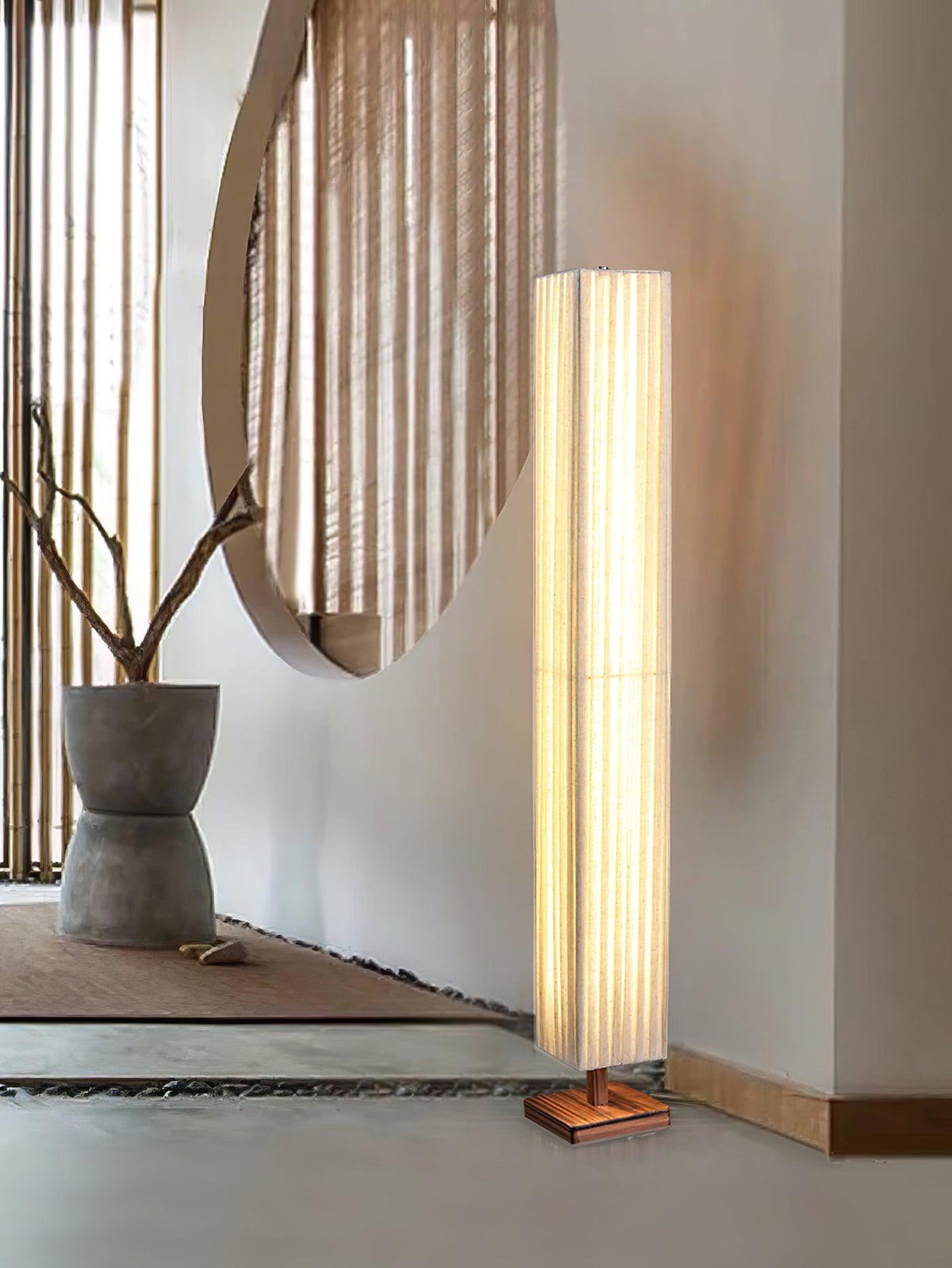 Clérachic floor lamp on a pale floor beside a round slatted wall panel, with a grey vase holding bare branches.