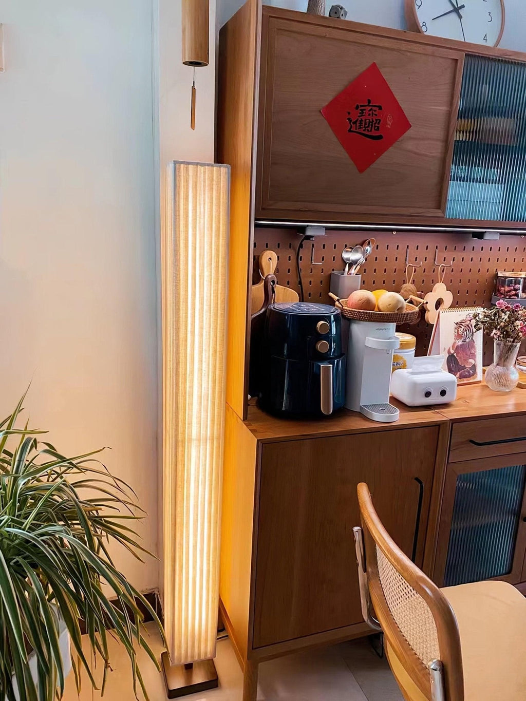 Close view of a Clérachic floor lamp beside a wooden kitchen dresser, with a rattan chair and a spider plant.