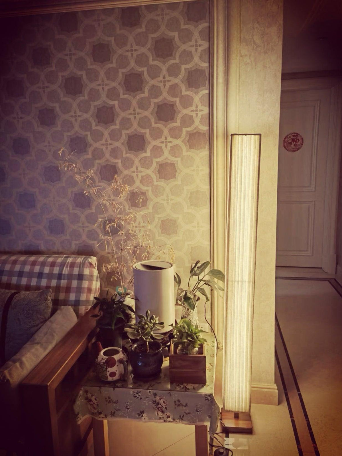 Clérachic floor lamp glowing beside a patterned wall and a table of small plants, in a warm, amber-toned room.