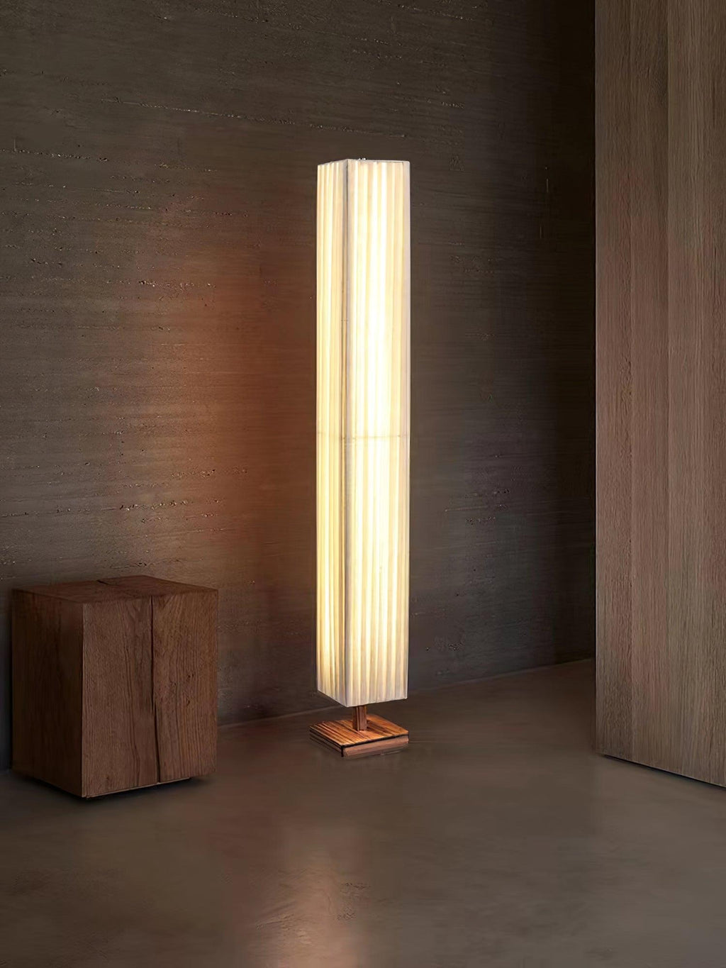Beige pleated Clérachic floor lamp standing on a concrete-look floor, with a block wooden stool beside it.