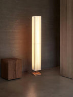 Beige pleated Clérachic floor lamp standing on a concrete-look floor, with a block wooden stool beside it.