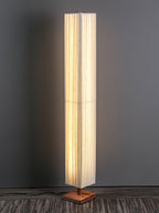 Lit Clérachic floor lamp on a square walnut base, with a cord trailing across a dark wood floor against a grey wall.