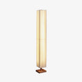 Tall pleated Clérachic floor lamp lit warm on a square wooden base, in a three-quarter view on a light grey backdrop.