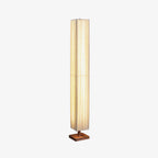 Tall pleated Clérachic floor lamp lit warm on a square wooden base, in a three-quarter view on a light grey backdrop.