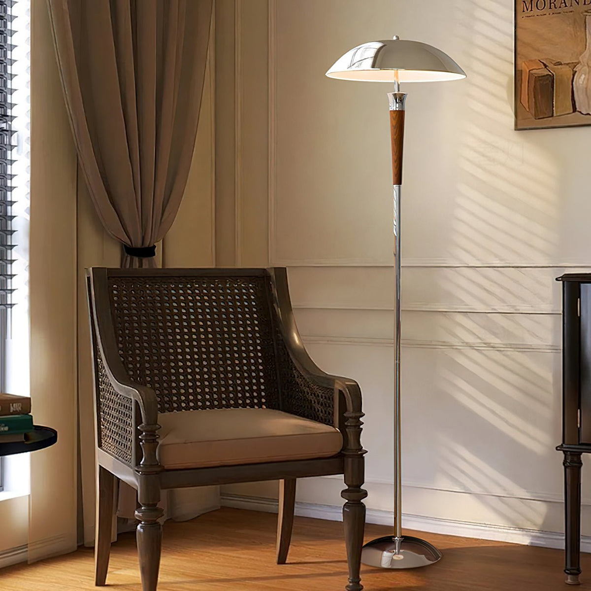 A Clérachic chrome-shaded floor lamp glows in a corner beside a dark rattan armchair, with a taupe curtain and panelled wall.