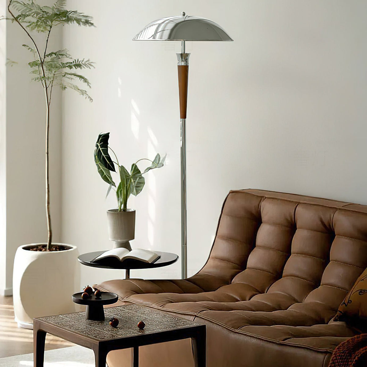 A tall Clérachic floor lamp with a chrome dome shade stands beside a tan leather sofa and a potted tree in a bright room.
