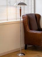 A Clérachic chrome-shaded floor lamp stands by a window with blinds, next to a brown leather wingback armchair on a herringbone floor.