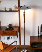 Side view of a Clérachic floor lamp with a chrome dome shade stands by a wooden desk, a tan leather chair and candlesticks.