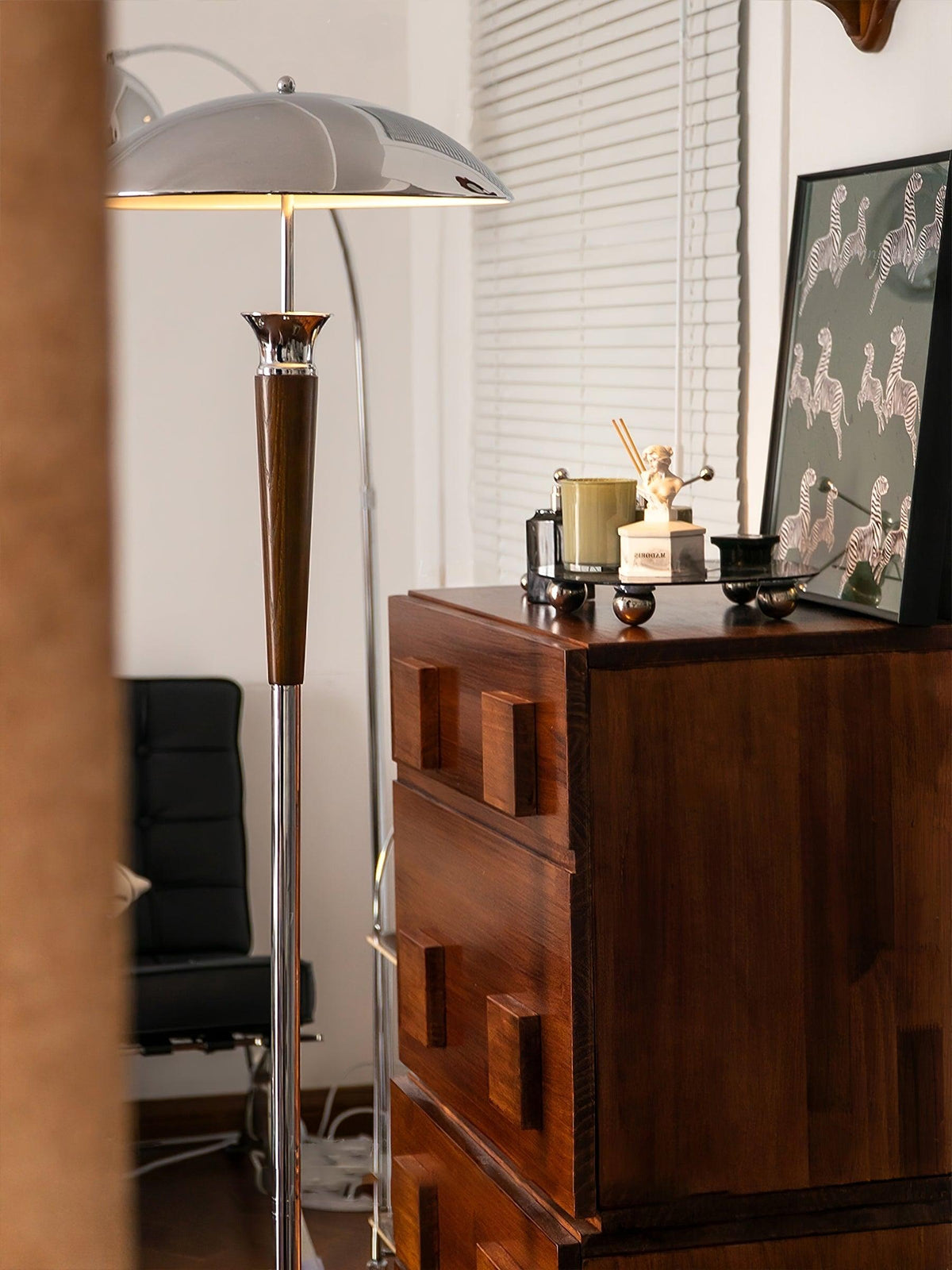 Lit Clérachic floor lamp with a chrome dome and walnut stem stands behind a dark wood chest, with a zebra-print wall hanging.