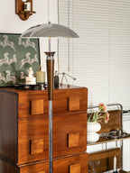 Slim Clérachic floor lamp with a chrome shade and walnut stem rises above a wood chest of drawers, with a patterned print and a window with blinds.