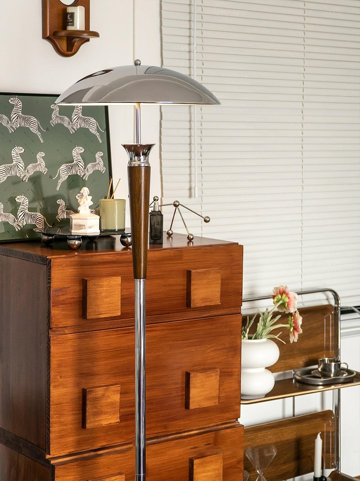 Slim Clérachic floor lamp with a chrome shade and walnut stem rises above a wood chest of drawers, with a patterned print and a window with blinds.