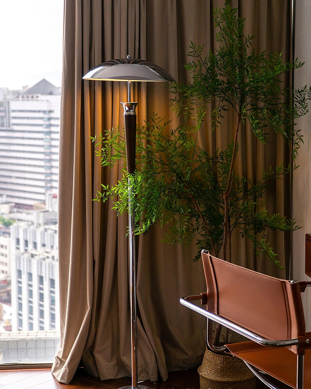 A Clérachic floor lamp in a living corner by tall brown curtains, with a leafy plant and a tan leather chair.