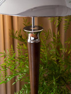 Close view of the chrome collar and walnut stem of this Clérachic floor lamp, with leafy branches and a curtain behind it.