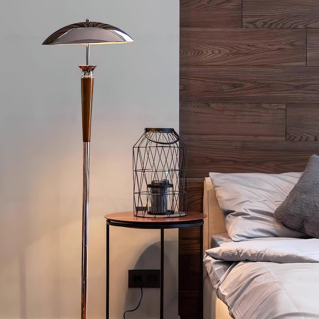 Clérachic floor lamp, shown with a chrome shade rises beside a dark wood headboard, a bedside table with a wire lantern and white bedding.