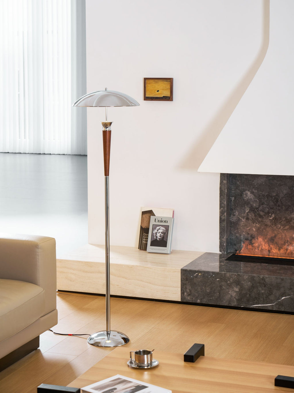 A Clérachic chrome-shaded floor lamp stands before a white fireplace with a dark stone hearth, beside a tan leather sofa.