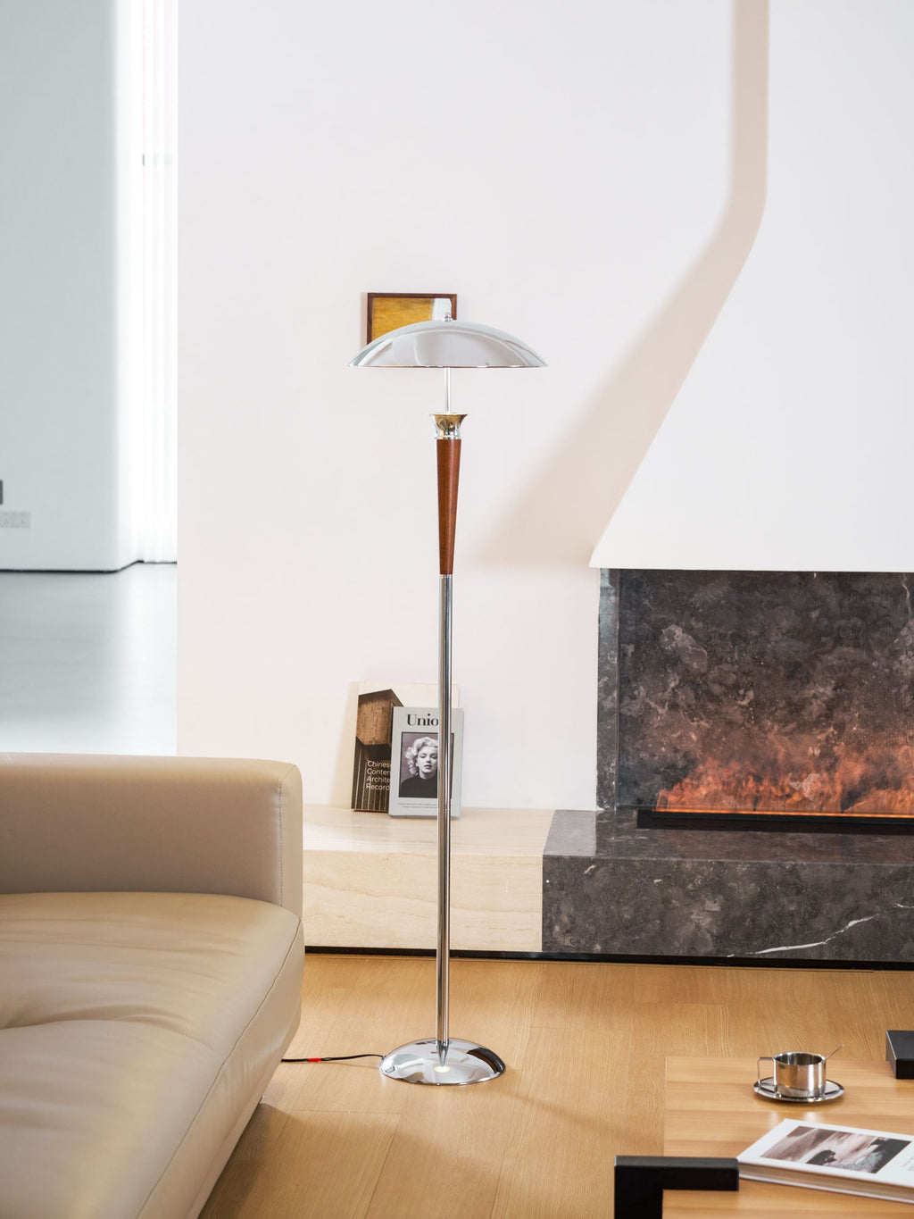 A Clérachic floor lamp stands in front of a stone fireplace, with a book and a small framed picture above the sofa arm.