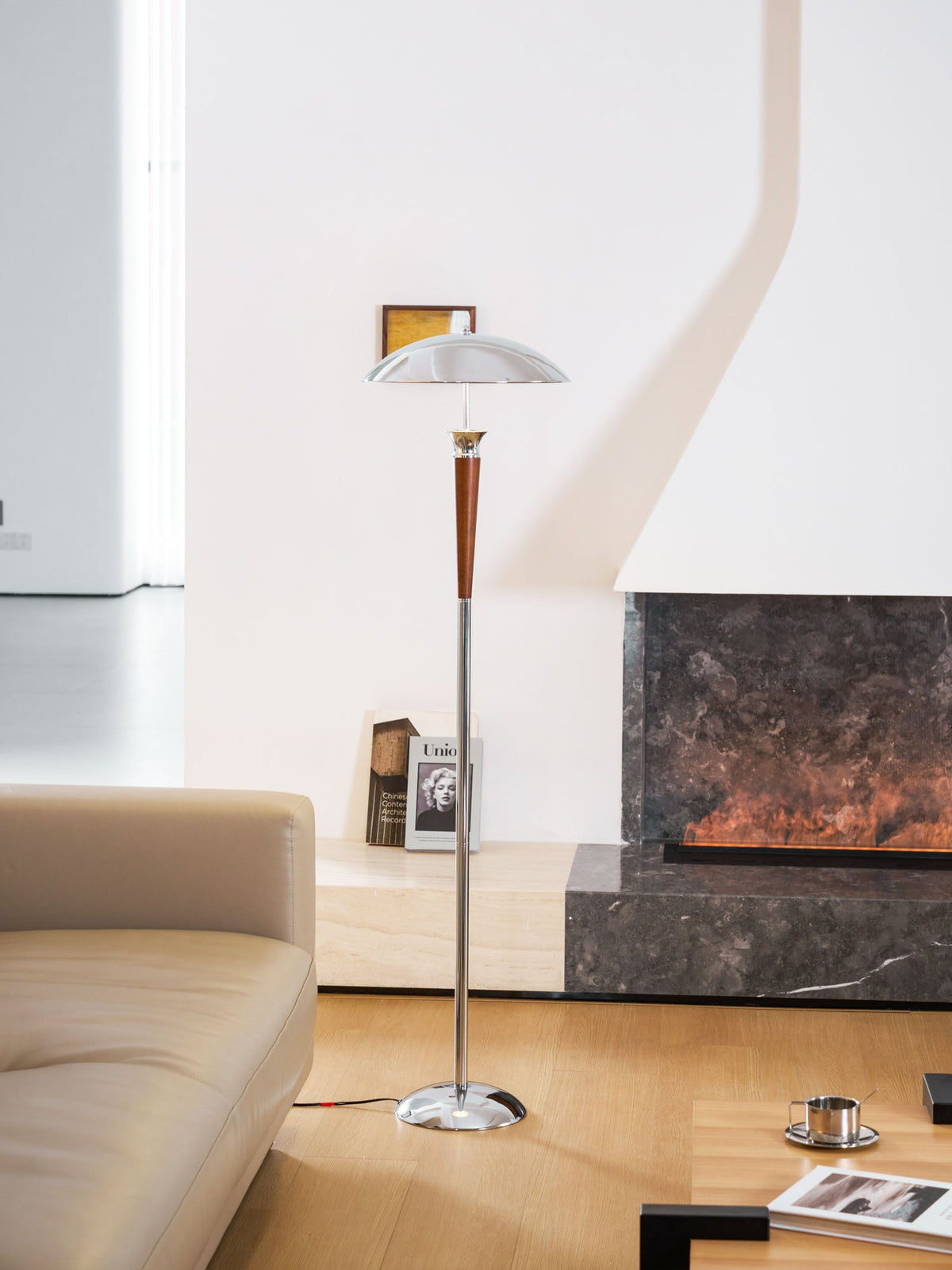 A Clérachic floor lamp stands in front of a stone fireplace, with a book and a small framed picture above the sofa arm.