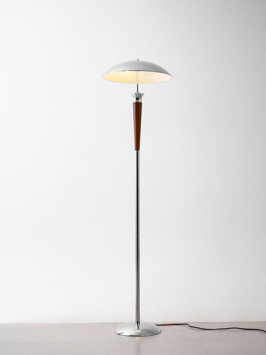Clérachic floor lamp beside furniture, with a chrome dome and walnut stem, standing on a grey floor with a dark cord, lit against a white wall.