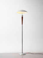 Clérachic floor lamp beside furniture, with a chrome dome and walnut stem, standing on a grey floor with a dark cord, lit against a white wall.