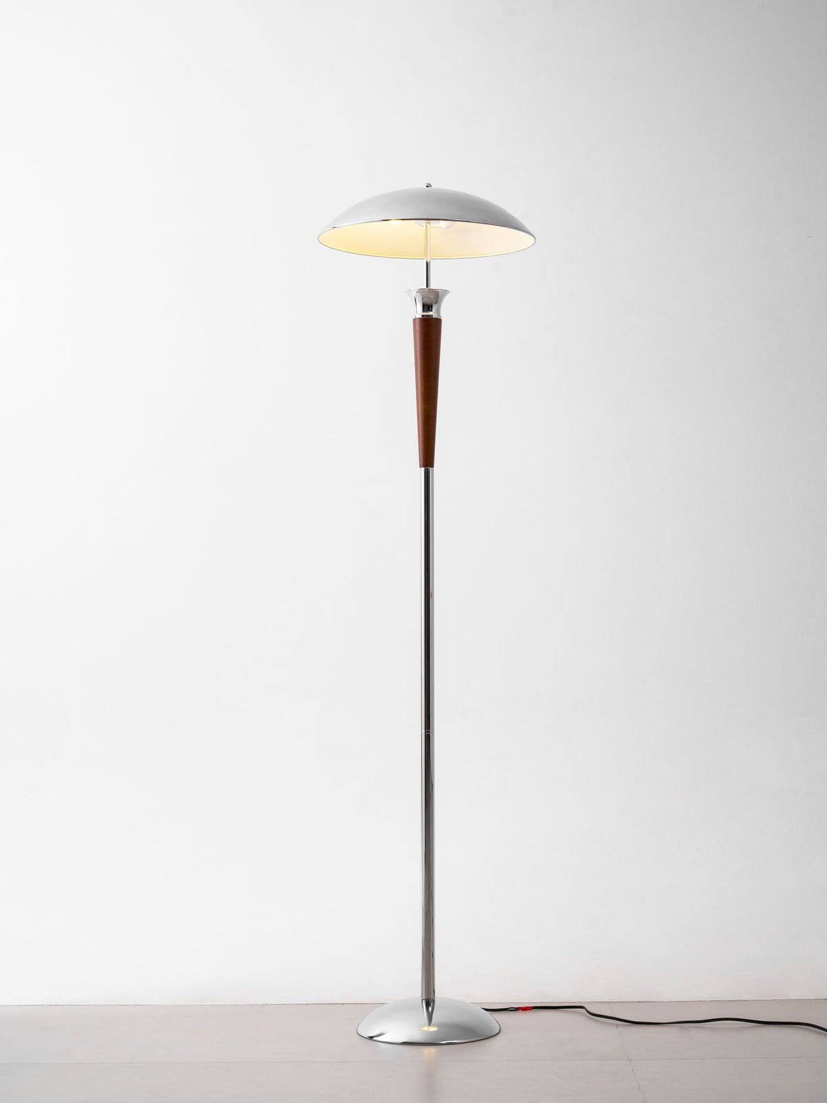 Clérachic floor lamp beside furniture, with a chrome dome and walnut stem, standing on a grey floor with a dark cord, lit against a white wall.