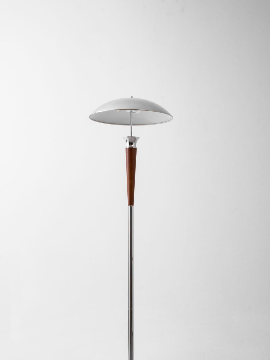 A Clérachic floor lamp with a chrome dome shade and walnut stem stands against a plain pale wall, shown at a distance.