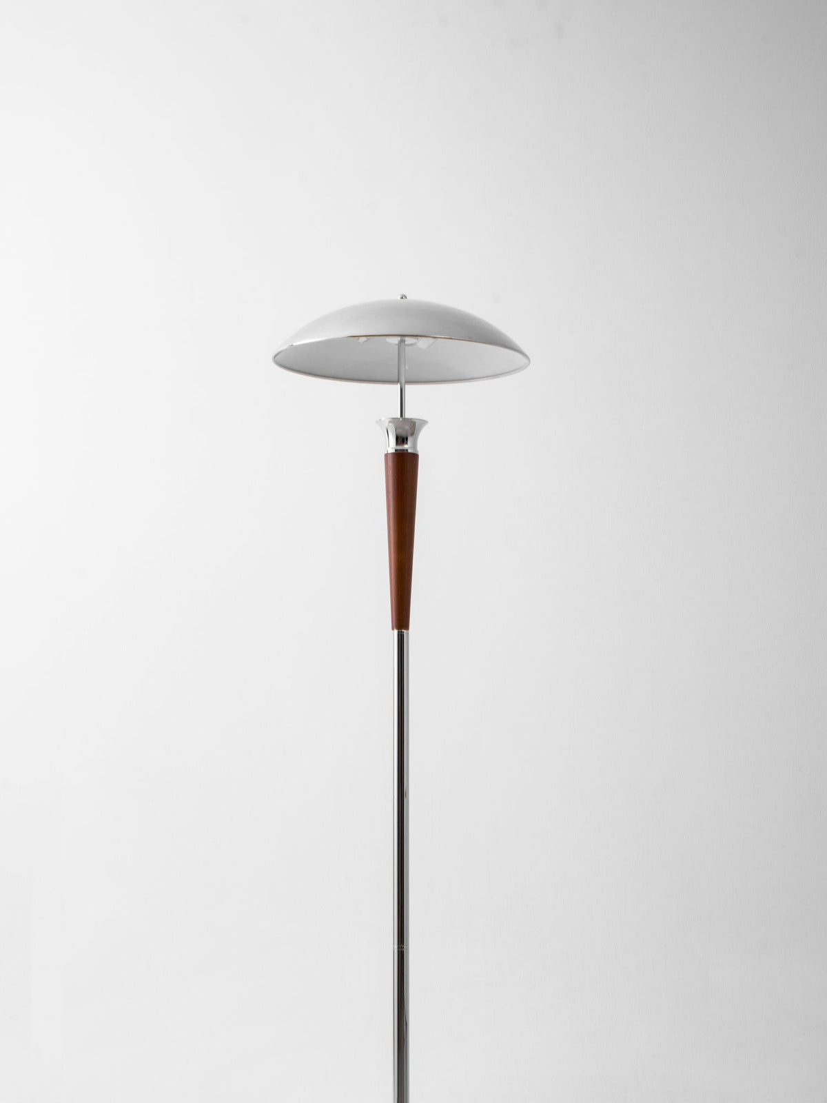 A Clérachic floor lamp with a chrome dome shade and walnut stem stands against a plain pale wall, shown at a distance.