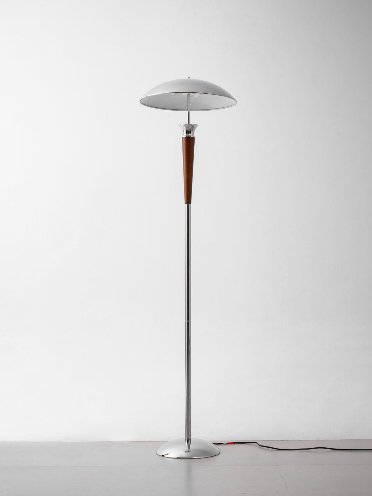 A Clérachic floor lamp with a chrome dome, walnut stem and round chrome base, standing on a grey floor against a white wall.