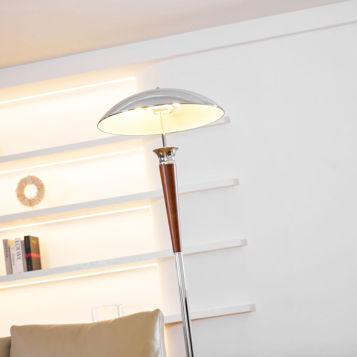 Low view up to the lit chrome shade of this Clérachic floor lamp, with glowing wall shelves and a sofa behind.