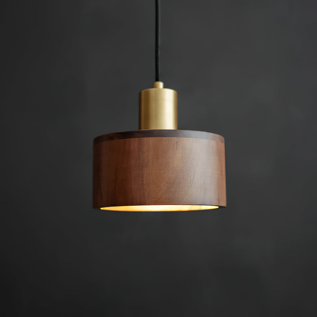 Front view of this Clérachic pendant with a brass cap and a walnut shade, its rim lit softly from below, on a dark backdrop.